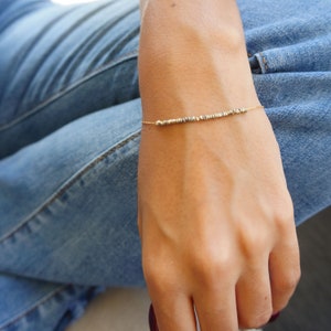 May include: A delicate gold chain bracelet with small silver beads. The bracelet is worn on a person's wrist.
