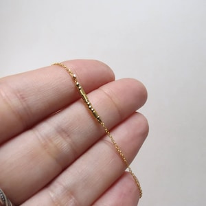 May include: Delicate gold necklace featuring a linear bar pendant with small, evenly spaced beads. The chain is fine and the pendant is a simple, elegant design. The necklace is shown against a neutral background.