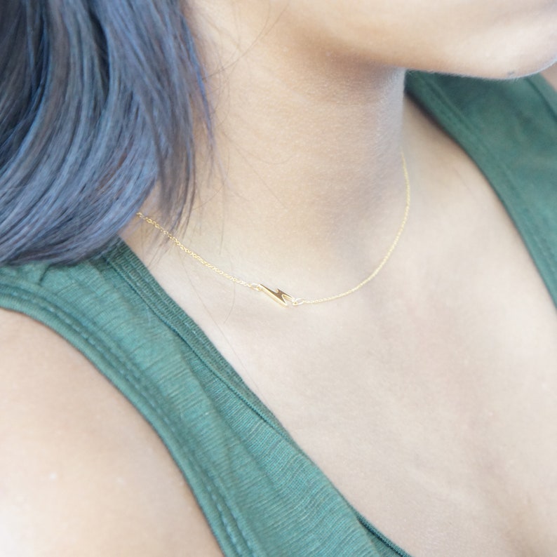 May include: A delicate gold necklace with a lightning bolt pendant. The necklace is worn on a person with dark hair and a green top. The pendant is small and minimalist, adding a touch of elegance to the neckline.