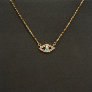 May include: A gold chain necklace with a small evil eye pendant. The pendant is decorated with clear rhinestones and has a blue center.