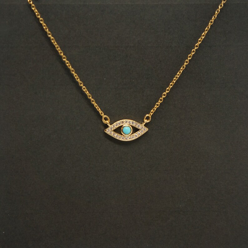 May include: A gold chain necklace with a small evil eye pendant. The pendant is decorated with clear rhinestones and has a blue center.