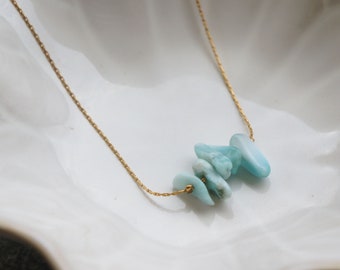 May include: A delicate gold chain necklace featuring a cluster of four small, polished, light blue larimar stone chips. The stones are irregularly shaped with smooth surfaces and are strung directly onto the thin, metallic gold-colored chain.