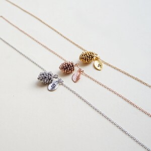 May include: Three necklaces featuring pine cone pendants and leaf charms in silver, rose gold, and gold. Each leaf charm has a different initial. The fine chains are displayed against a cream background. The necklaces are a nature-inspired design.