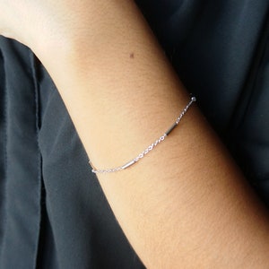 May include: A delicate silver bracelet featuring a fine chain with two small, horizontal bar accents. The bracelet is shown on a person's arm, against a dark background. The minimalist design suggests a focus on simplicity and elegance.