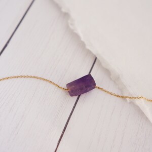 May include: A delicate gold chain necklace with a single, raw amethyst stone pendant. The stone is a light purple color and has a natural, irregular shape.