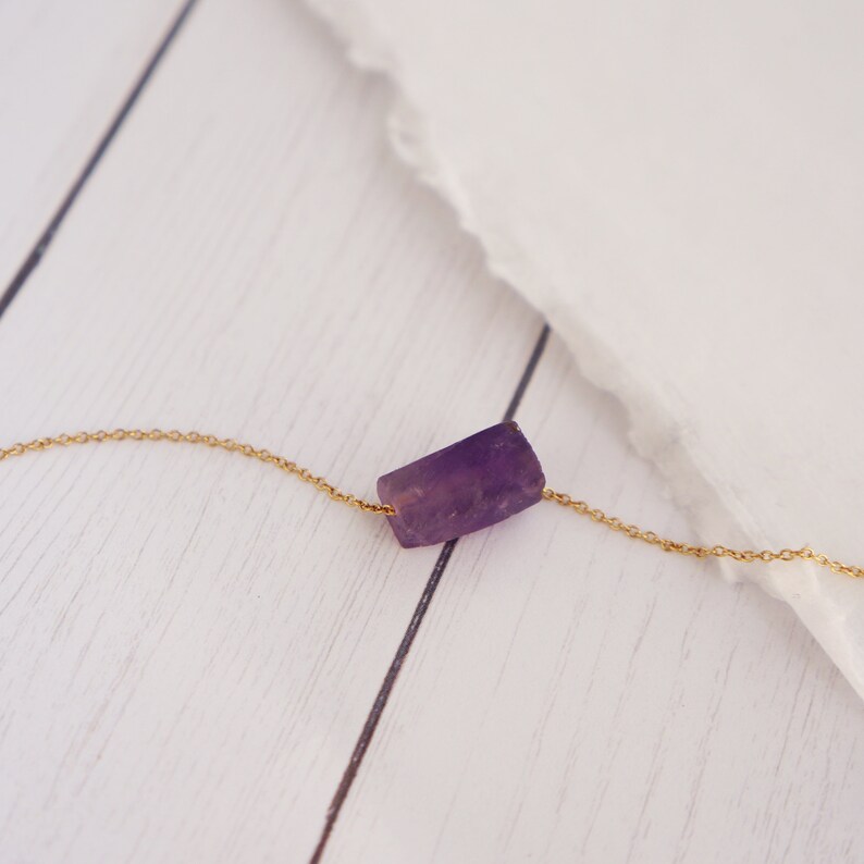 May include: A delicate gold chain necklace with a single, raw amethyst stone pendant. The stone is a light purple color and has a natural, irregular shape.