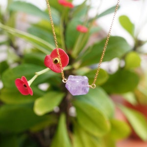 Puede incluir: Un collar de cadena dorada con un colgante de cristal de amatista p&uacute;rpura. El collar est&aacute; colgado de una planta verde con flores rojas.