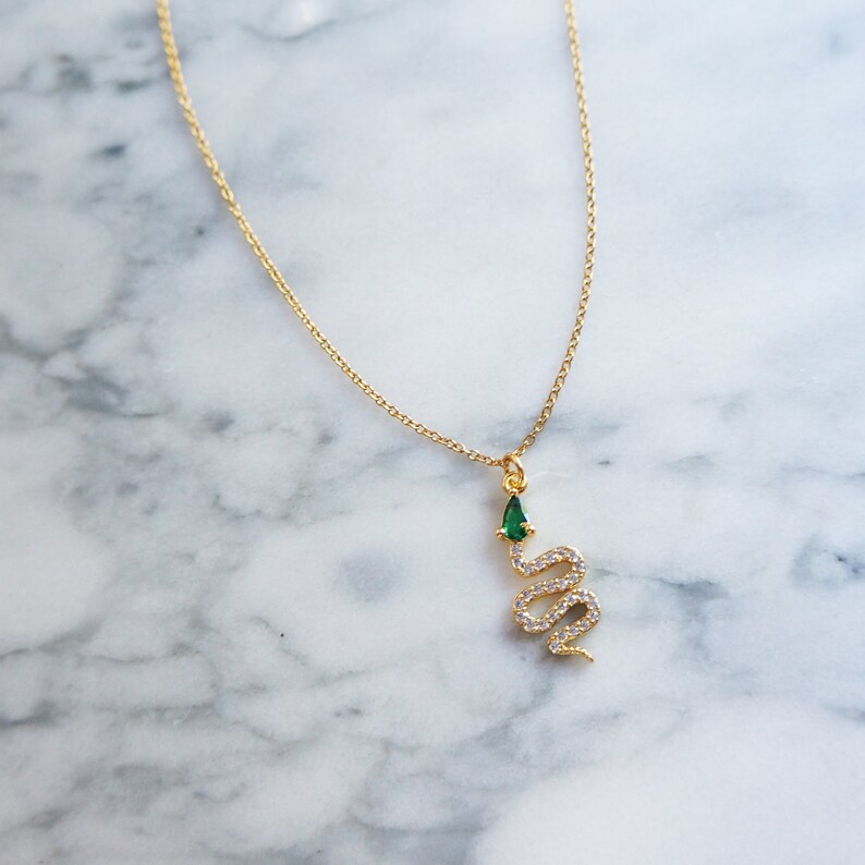 May include: A gold chain necklace with a snake pendant. The snake is encrusted with clear rhinestones and has a green gemstone on its head.