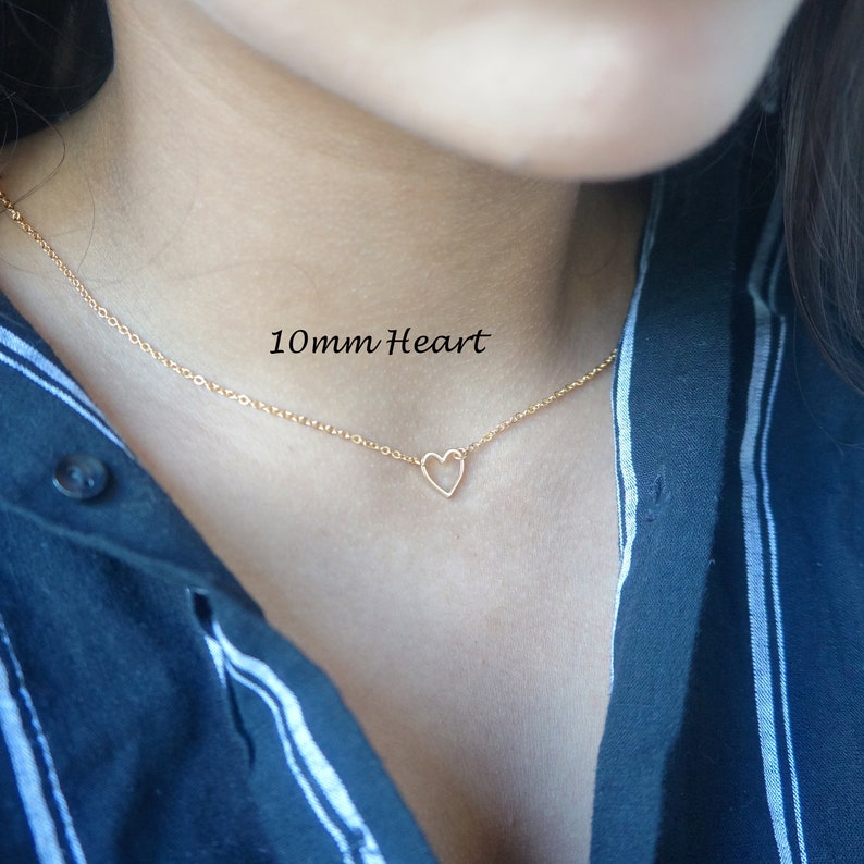 Puede incluir: Un collar de cadena dorada con un colgante de coraz&oacute;n abierto de 10 mm. El collar lo lleva una persona con una camisa a rayas azules y blancas.