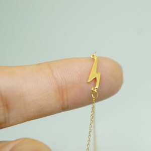 May include: A gold-colored necklace featuring a lightning bolt pendant. The pendant is a stylized zig-zag shape, attached to a delicate chain. The necklace is shown against a light background.