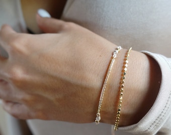May include: Two gold bracelets on a person's wrist. The top bracelet is a delicate chain with small, round beads. The bottom bracelet is a thin, flat band with small, clear stones.