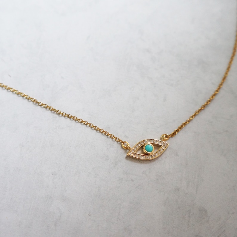 May include: A gold chain necklace with a turquoise evil eye pendant. The pendant is surrounded by small clear stones.