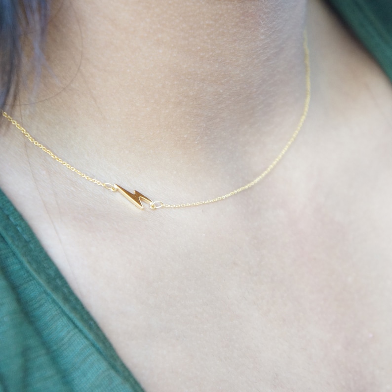 May include: A delicate gold-colored necklace featuring a lightning bolt pendant. The necklace is shown against a skin tone background and a green garment. The lightning bolt is a simple, minimalist design.