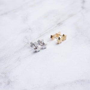 K&ouml;nnte beinhalten: Zwei Paar sternf&ouml;rmige Ohrstecker. Ein Paar ist silberfarben, das andere goldfarben. Die Ohrringe liegen auf einer wei&szlig;en Marmoroberfl&auml;che. Die Ohrringe sind klein und zart, mit einem schlichten Design.