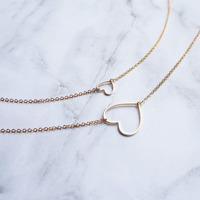 Puede incluir: Un collar de oro con dos delicados colgantes de coraz&oacute;n abierto. El colgante de coraz&oacute;n m&aacute;s grande est&aacute; en la parte inferior del collar, y el colgante de coraz&oacute;n m&aacute;s peque&ntilde;o est&aacute; m&aacute;s arriba.