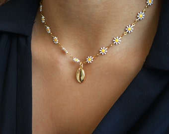 May include: A gold-colored necklace featuring a daisy chain design with white and yellow enamel flowers. A gold seashell pendant hangs from the center. The necklace is worn against a dark blue garment.