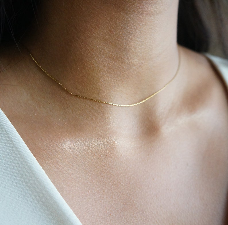 May include: A delicate gold necklace resting on a person's neck. The necklace is a thin chain with a subtle, shimmering appearance. The background is blurred, focusing attention on the jewelry.