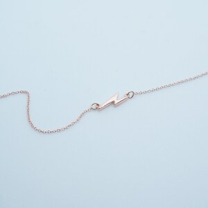 May include: A delicate rose gold necklace featuring a lightning bolt pendant. The pendant is a stylized zig-zag shape, connected to a fine chain. The necklace is displayed against a light blue background, highlighting the jewelry's minimalist design.