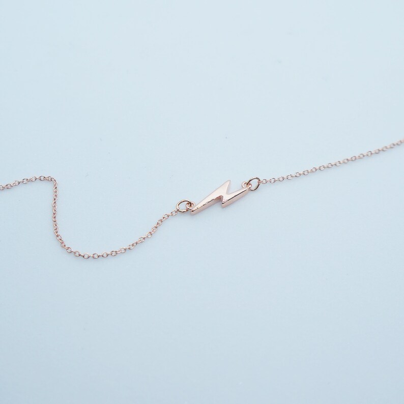 May include: A delicate rose gold necklace featuring a lightning bolt pendant. The pendant is a stylized zig-zag shape, connected to a fine chain. The necklace is displayed against a light blue background, highlighting the jewelry's minimalist design.