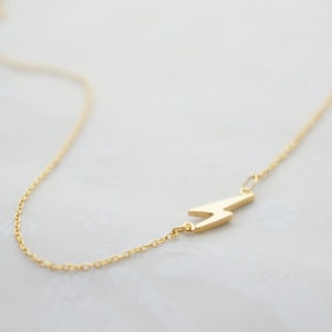 May include: A gold-colored necklace featuring a lightning bolt pendant. The pendant is a stylized zig-zag shape, attached to a delicate chain. The necklace is displayed on a light-colored surface.