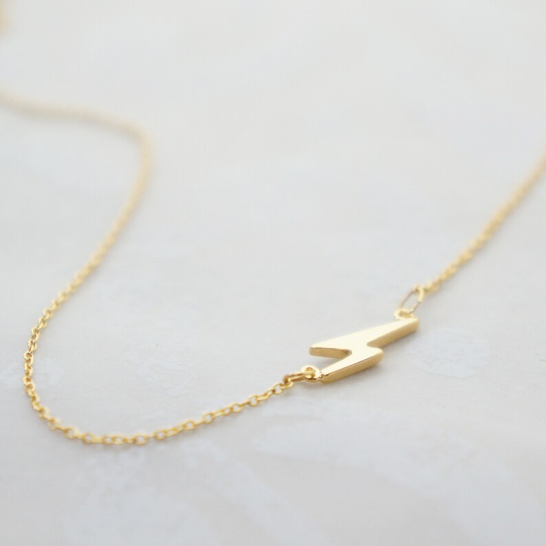 May include: A gold-colored necklace featuring a lightning bolt pendant. The pendant is a stylized zig-zag shape, attached to a delicate chain. The necklace is displayed on a light-colored surface.