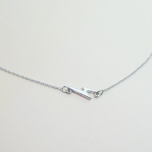 May include: A silver-colored necklace featuring a lightning bolt pendant. The pendant is a stylized zig-zag shape, connected to a delicate chain. The necklace is displayed against a light background, highlighting the jewelry's design.