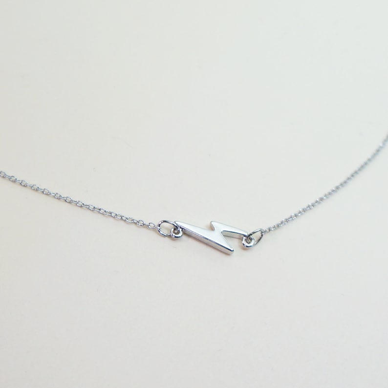 May include: A silver-colored necklace featuring a lightning bolt pendant. The pendant is a stylized zig-zag shape, connected to a delicate chain. The necklace is displayed against a light background, highlighting the jewelry's design.