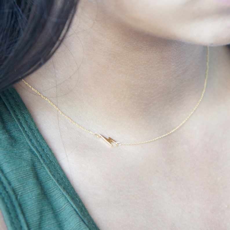 May include: Delicate gold-colored necklace featuring a lightning bolt pendant. The pendant is small and positioned to the side of the chain. The necklace is worn against a green top.