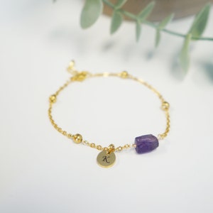 May include: A gold chain bracelet with a small gold charm that says "K" and a purple amethyst stone.