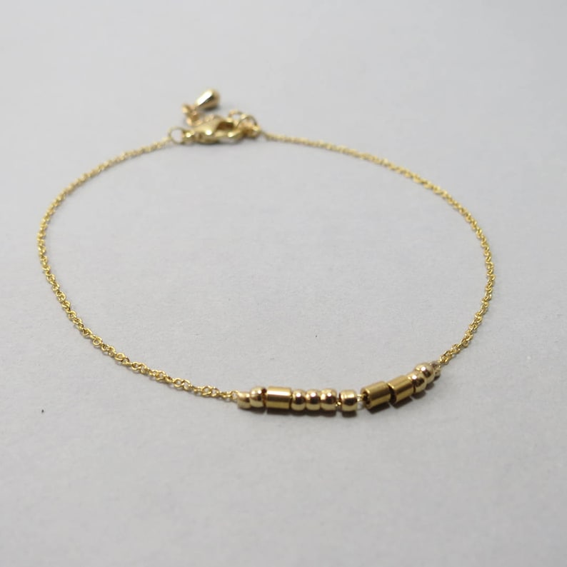 May include: Delicate gold-toned bracelet featuring a series of small, cylindrical and spherical beads. The bracelet has a fine chain and an adjustable clasp. The jewelry is displayed on a neutral gray background, highlighting the gold color.