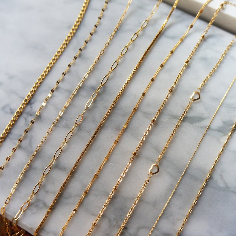 May include: A collection of delicate gold necklaces displayed on a marble surface. The necklaces feature various chain styles, including box, paperclip, and beaded designs. Some necklaces have heart-shaped pendants.