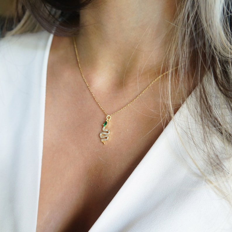May include: A gold chain necklace with a small snake pendant. The snake is adorned with clear rhinestones and has a green gemstone for an eye.