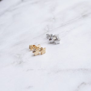 K&ouml;nnte beinhalten: Zwei Paar sternf&ouml;rmige Ohrstecker. Ein Paar ist goldfarben, das andere silberfarben. Die Ohrringe sind klein und zart, mit einem einfachen, minimalistischen Design. Die Ohrringe werden auf einer wei&szlig;en Marmoroberfl&auml;che pr&auml;sentiert.