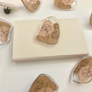 May include: A set of eight clear resin pins featuring abstract line art faces with brown and beige accents. The pins are displayed on a white surface.