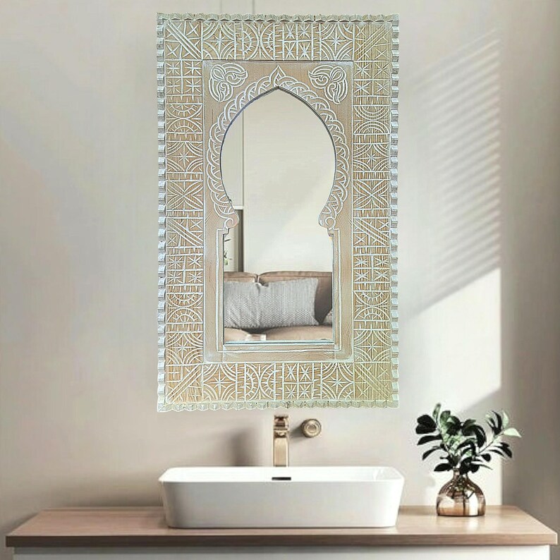 May include: A white wooden mirror with an intricate carved design. The mirror has an arched top and a rectangular frame. The mirror is hanging on a white wall above a white sink.