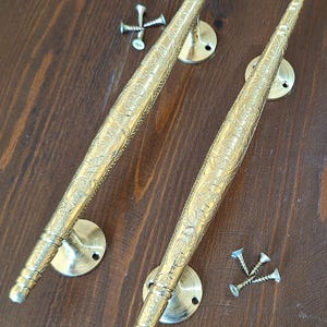 Puede incluir: Dos tiradores de puerta dorados con intrincados diseños florales. Los tiradores son largos y delgados con un extremo redondeado. Están unidos a placas redondas de metal con tornillos.