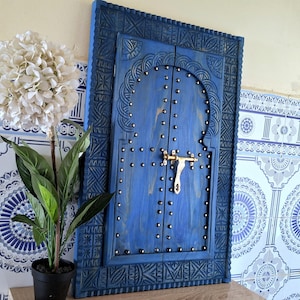 Puede incluir: Una puerta de madera azul con tallas intrincadas y un pestillo dorado. La puerta está enmarcada por un marco de madera tallada y se coloca sobre un fondo de azulejos blancos.
