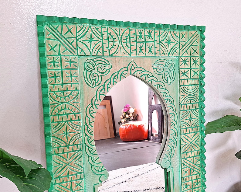 May include: A green wooden mirror with an intricate carved design. The mirror has a traditional Moroccan style arch and is framed with a decorative border.