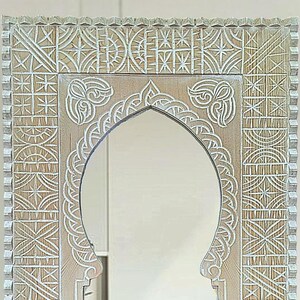 May include: A white wooden mirror with an ornate carved frame. The frame features a geometric pattern with a central arch.