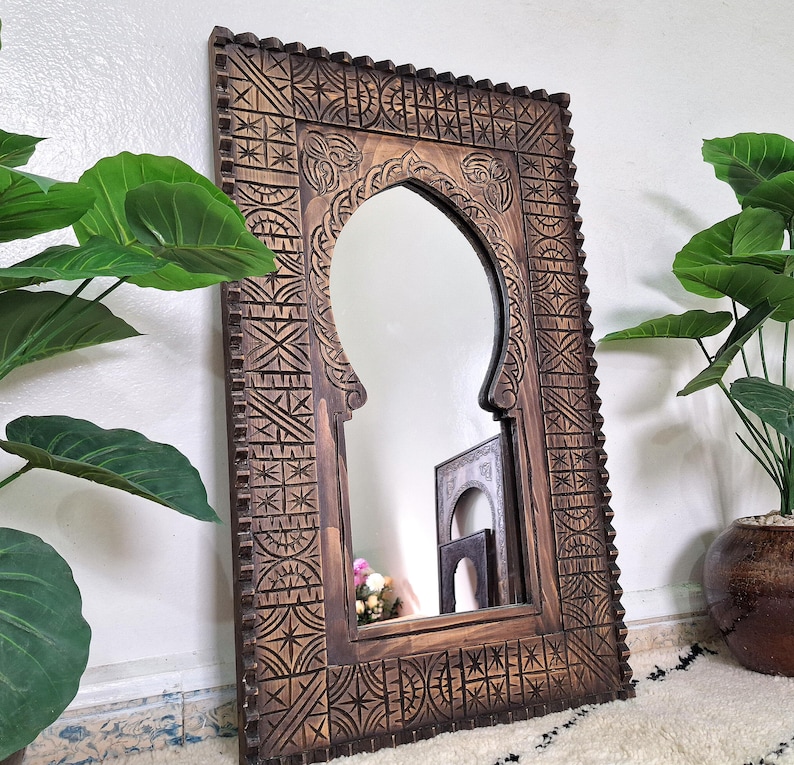 May include: A carved wooden mirror with an arched opening and intricate geometric patterns. The mirror has a dark brown finish and is framed by a thick, ornate border.