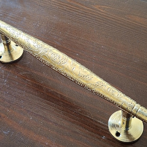 May include: A gold-toned door handle with an ornate design. The handle is long and has a decorative pattern on the surface.
