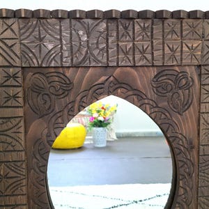 May include: A dark brown wooden mirror with a carved frame. The frame has intricate designs and patterns. The mirror is oval shaped and has a beveled edge.