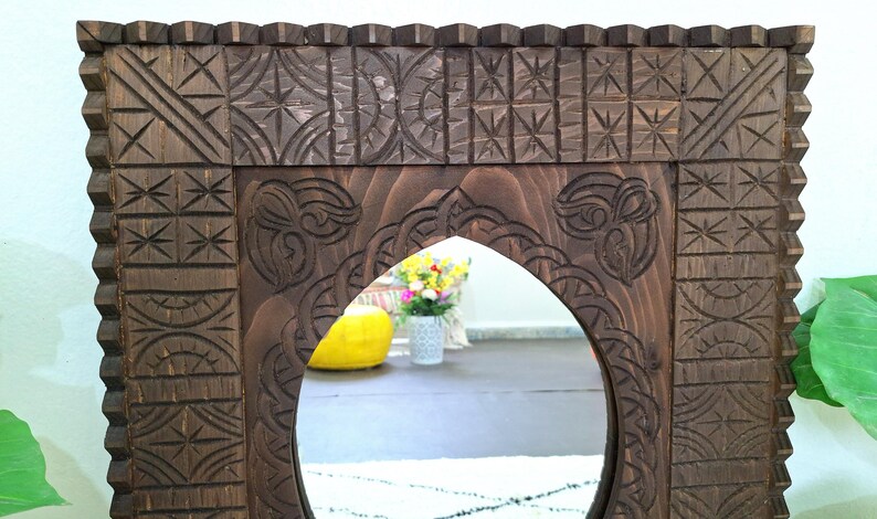 May include: A dark brown wooden mirror with a carved frame. The frame has intricate designs and patterns. The mirror is oval shaped and has a beveled edge.