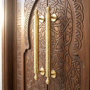 May include: A detailed view of a wooden door featuring elaborate carved designs and two long, gold-coloured door handles. The handles are cylindrical with decorative patterns and circular mounting plates. The wood of the door has a warm, brown hue.