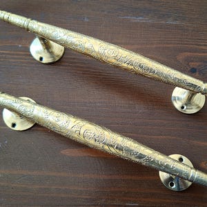 Könnte beinhalten: Zwei verzierte Türklinken aus Messing mit aufwendigen floralen Mustern. Die Griffe sind lang und schlank mit abgerundetem Ende.