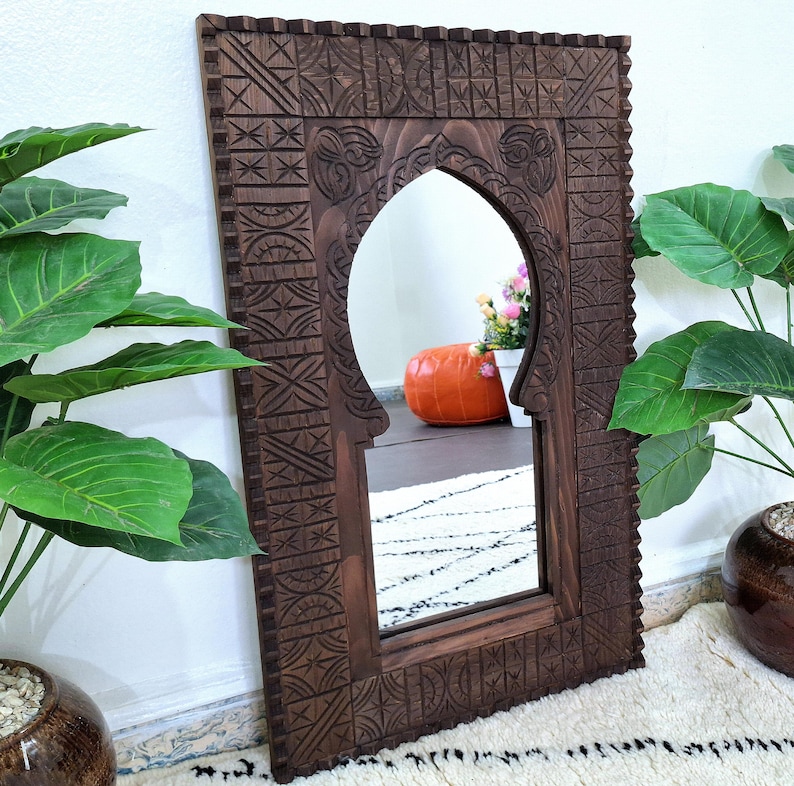 May include: A dark brown wooden mirror with an intricately carved frame. The mirror has an arched top and a Moroccan-style design.