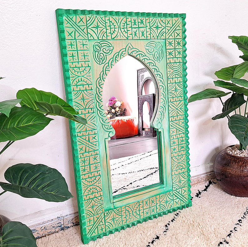 May include: A green wooden mirror with intricate carved designs. The mirror has an arched top and is framed by a border of geometric patterns.
