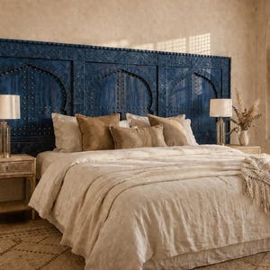 Può includere: Camera da letto con un letto grande con testiera blu intagliata. Il letto è coperto da un piumino e cuscini color crema. Due comodini con lampade fiancheggiano il letto. Un tappeto decorativo e una grande pianta completano la scena.