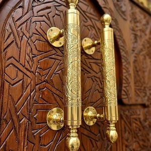 Può includere: Primo piano di una porta in legno con intagli intricati e due maniglie dorate ornate. Le maniglie presentano dettagliati motivi floreali e sono montate su placche rotonde dorate. La porta è di un marrone caldo e ricco.