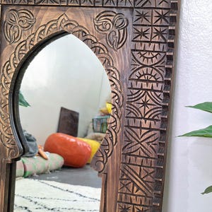 May include: A wooden mirror with an intricately carved frame. The frame features a Moroccan-inspired design with geometric patterns and an arched opening. The mirror is reflecting a room with a rug and furniture.